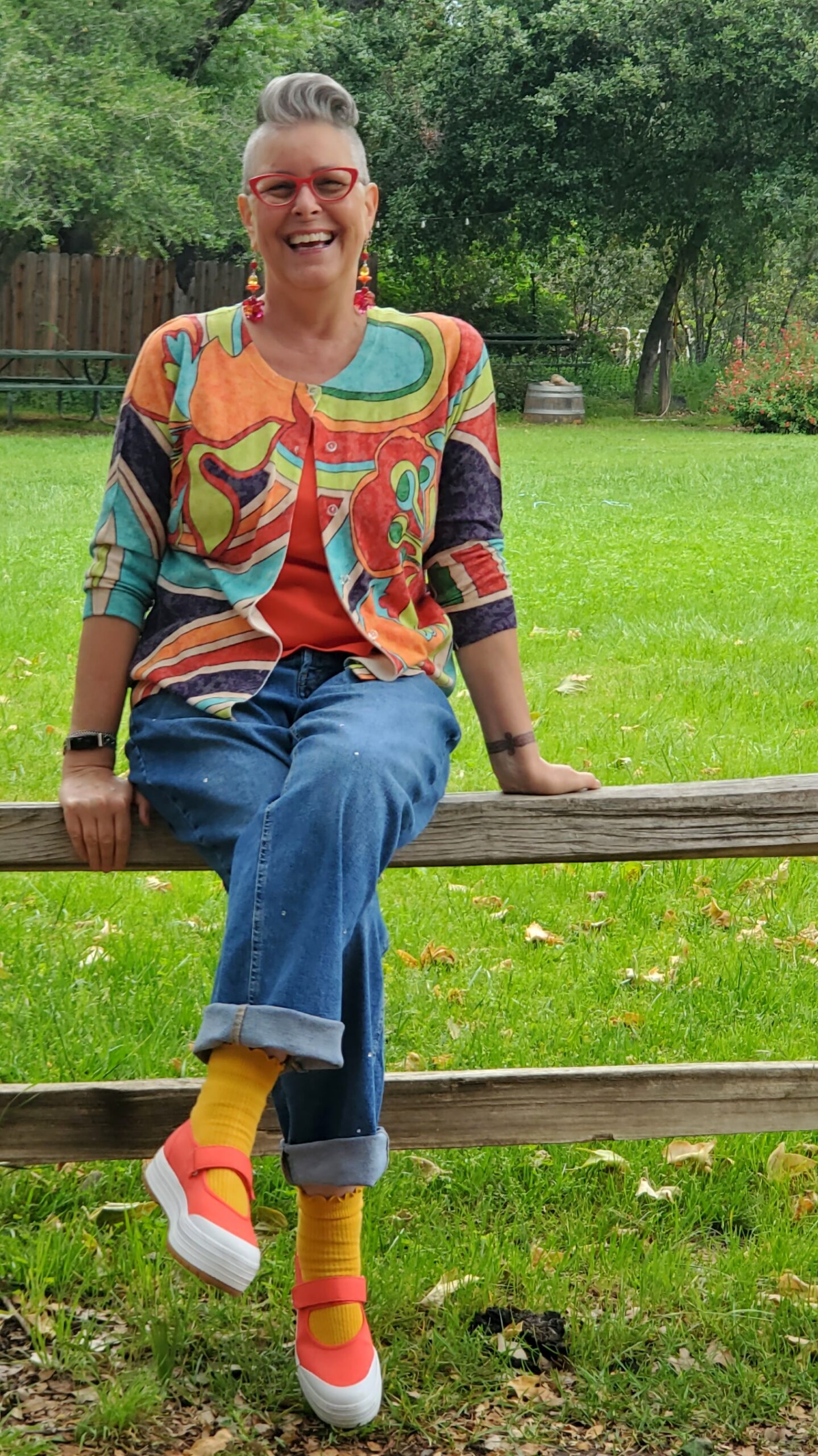 Lisa sitting on a fence in a colorful sweater, jeans, orange shoes and bright green grass and trees in the background.